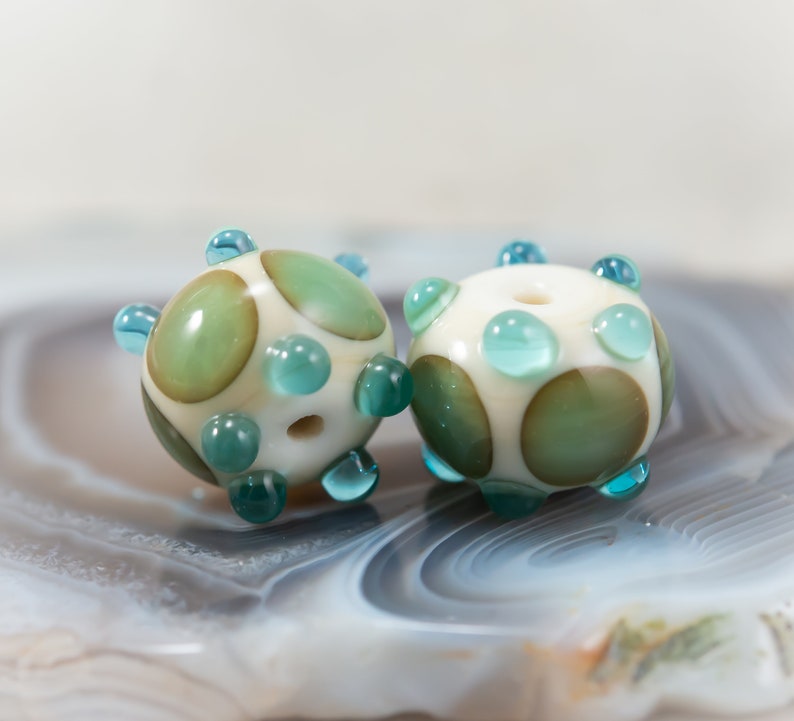 May include: Two handmade glass beads. Each bead is spherical, with a creamy white base color and large, irregular green and brown spots. Small, turquoise-colored glass accents are evenly spaced around the bead's surface. Each bead has a hole for stringing.