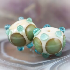 May include: Two handmade glass beads. Each bead is round with a cream-colored base, olive green and white polka dots, and light blue accents. The beads have a hole through the center, and are suitable for jewelry making.