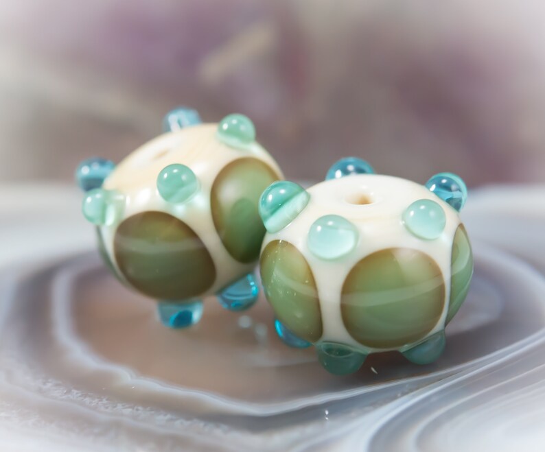 May include: Two handmade glass beads. Each bead is round with a cream-colored base, olive green and white polka dots, and light blue accents. The beads have a hole through the center, and are suitable for jewelry making.