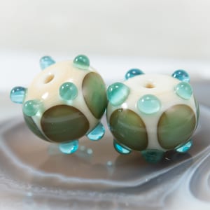 May include: Two handmade glass beads. Each bead is round with a cream-colored base, green and brown spots, and turquoise accents. The beads have a hole through the center for stringing. The beads are displayed on a light gray surface.