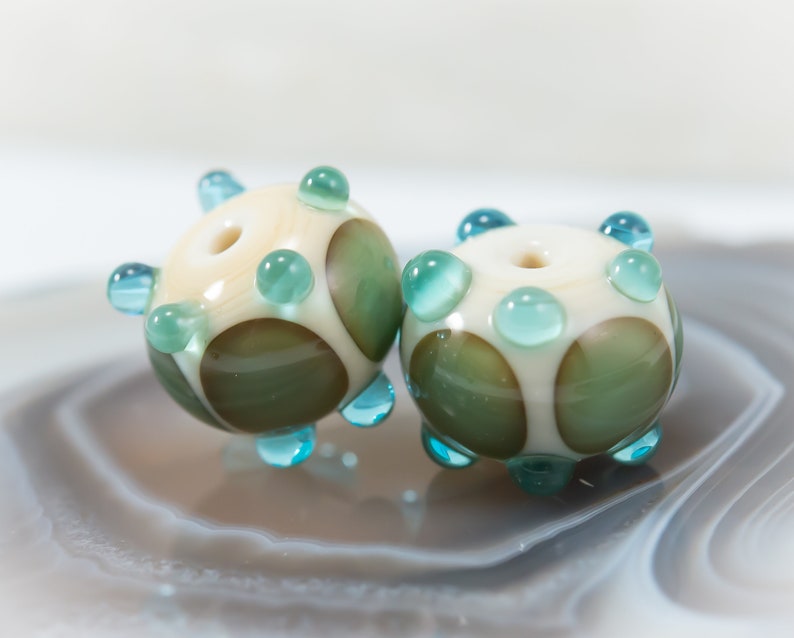 May include: Two handmade glass beads. Each bead is round with a cream-colored base, green and brown spots, and turquoise accents. The beads have a hole through the center for stringing. The beads are displayed on a light gray surface.
