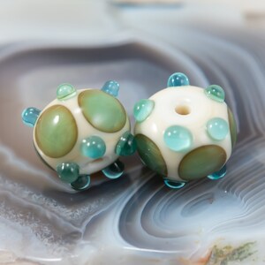 May include: Two handmade glass beads. Each bead is round with a cream-colored base, adorned with green and teal accents. The beads have a spotted pattern and small teal protrusions. The beads are likely for jewelry making.