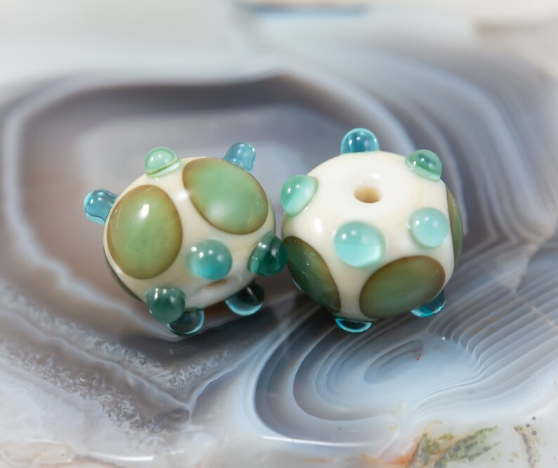 May include: Two handmade glass beads. Each bead is round with a cream-colored base, adorned with green and teal accents. The beads have a spotted pattern and small teal protrusions. The beads are likely for jewelry making.