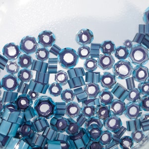 May include: An assortment of blue and purple faceted beads in various shapes, including round, octagonal, and cylindrical. The beads are scattered, creating a textured and visually appealing display. The beads are likely for jewelry making or crafts.