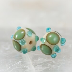 May include: Two handmade glass beads. Each bead is spherical, with a cream-colored base and large, olive-green and brown-toned spots. Small, light-blue glass dots are evenly spaced around the bead's surface. A hole runs through the center of each bead.