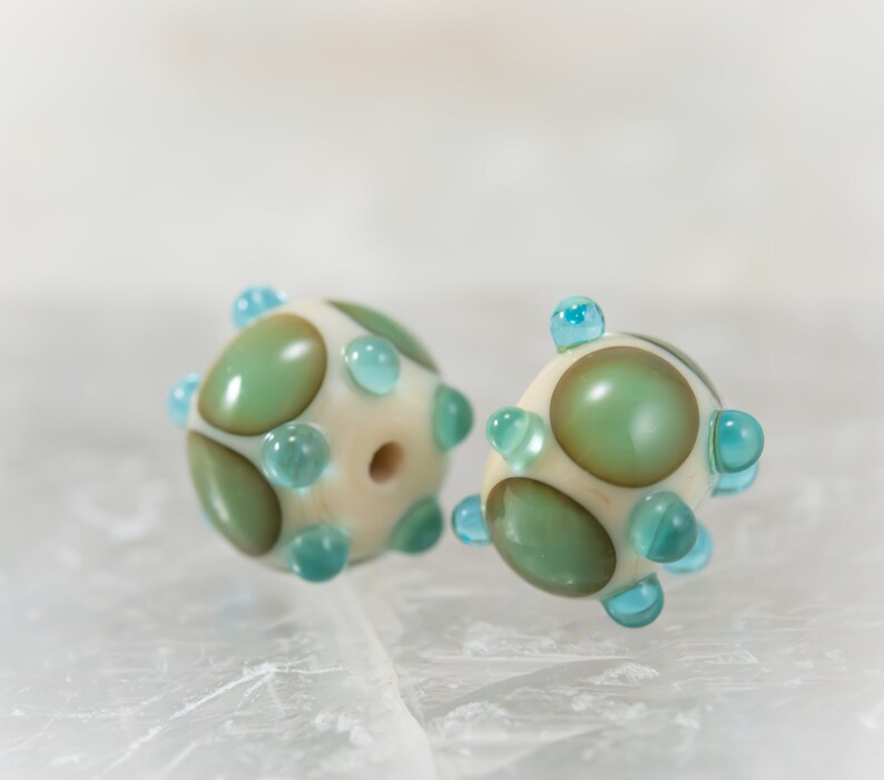 May include: Two handmade glass beads. Each bead is spherical, with a cream-colored base and large, olive-green and brown-toned spots. Small, light-blue glass dots are evenly spaced around the bead's surface. A hole runs through the center of each bead.