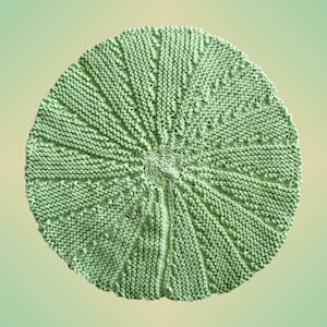 Könnte beinhalten: Ein runder, gestrickter Hut in hellgrüner Farbe. Der Hut hat ein strukturiertes Muster, das von der Mitte ausstrahlt, mit einem Spiraldesign am Oberteil. Der Strickstoff wirkt weich und flexibel, geeignet für Kopfbedeckungen.