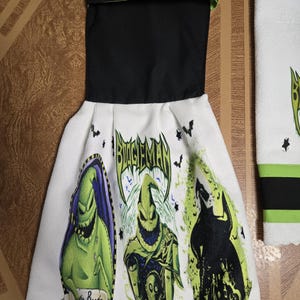 Puede incluir: Toalla de cocina blanca con parte superior negra y ribete verde. La toalla presenta tres im&aacute;genes del personaje Oogie Boogie de The Nightmare Before Christmas, con el texto "Boogie Man" y gr&aacute;ficos de murci&eacute;lagos.
