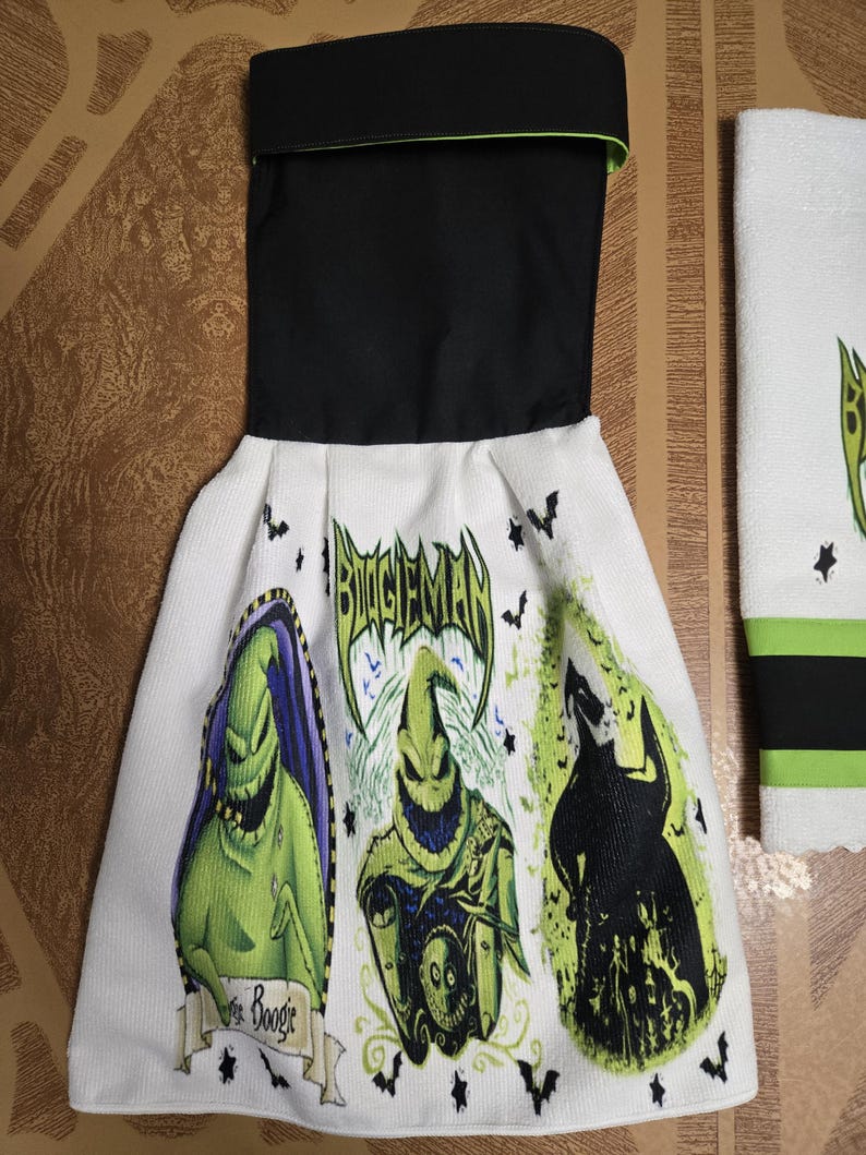 Puede incluir: Toalla de cocina blanca con parte superior negra y ribete verde. La toalla presenta tres im&aacute;genes del personaje Oogie Boogie de The Nightmare Before Christmas, con el texto "Boogie Man" y gr&aacute;ficos de murci&eacute;lagos.