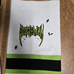 Puede incluir: Toalla de mano blanca con la palabra "BOOGIEMAN" en letras verdes y negras. La toalla tiene una franja verde y negra en la parte inferior, con gr&aacute;ficos de murci&eacute;lagos y estrellas.
