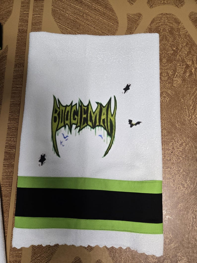 Puede incluir: Toalla de mano blanca con la palabra "BOOGIEMAN" en letras verdes y negras. La toalla tiene una franja verde y negra en la parte inferior, con gr&aacute;ficos de murci&eacute;lagos y estrellas.