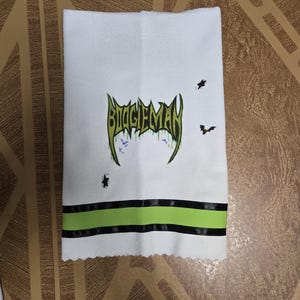 Puede incluir: Toalla de mano blanca con el texto "BIGGIEMAN" en verde y negro, junto con gr&aacute;ficos de murci&eacute;lagos y estrellas. La toalla tiene una franja verde y negra en la parte inferior.