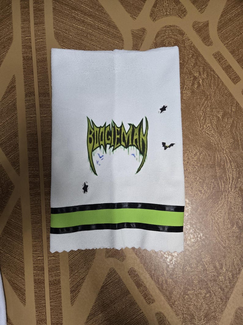 Puede incluir: Toalla de mano blanca con el texto "BIGGIEMAN" en verde y negro, junto con gr&aacute;ficos de murci&eacute;lagos y estrellas. La toalla tiene una franja verde y negra en la parte inferior.