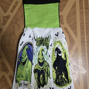 Puede incluir: Toalla de cocina blanca con tres im&aacute;genes de Oogie Boogie de The Nightmare Before Christmas. La toalla tiene una parte superior negra y verde con las palabras "Oogie Boogie" y gr&aacute;ficos de murci&eacute;lagos.