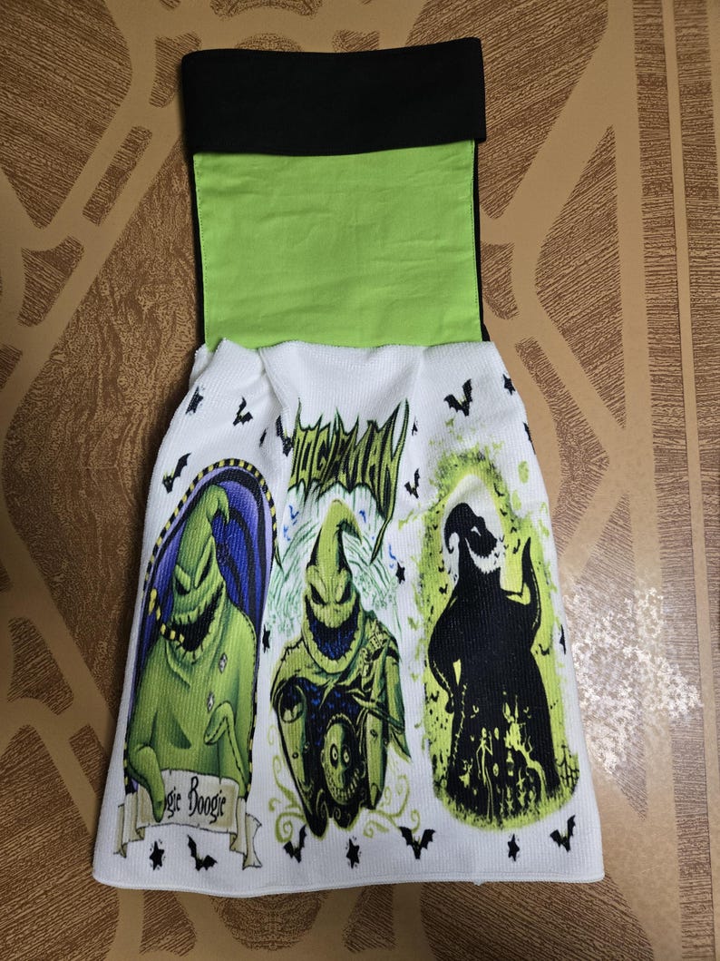 Puede incluir: Toalla de cocina blanca con tres im&aacute;genes de Oogie Boogie de The Nightmare Before Christmas. La toalla tiene una parte superior negra y verde con las palabras "Oogie Boogie" y gr&aacute;ficos de murci&eacute;lagos.