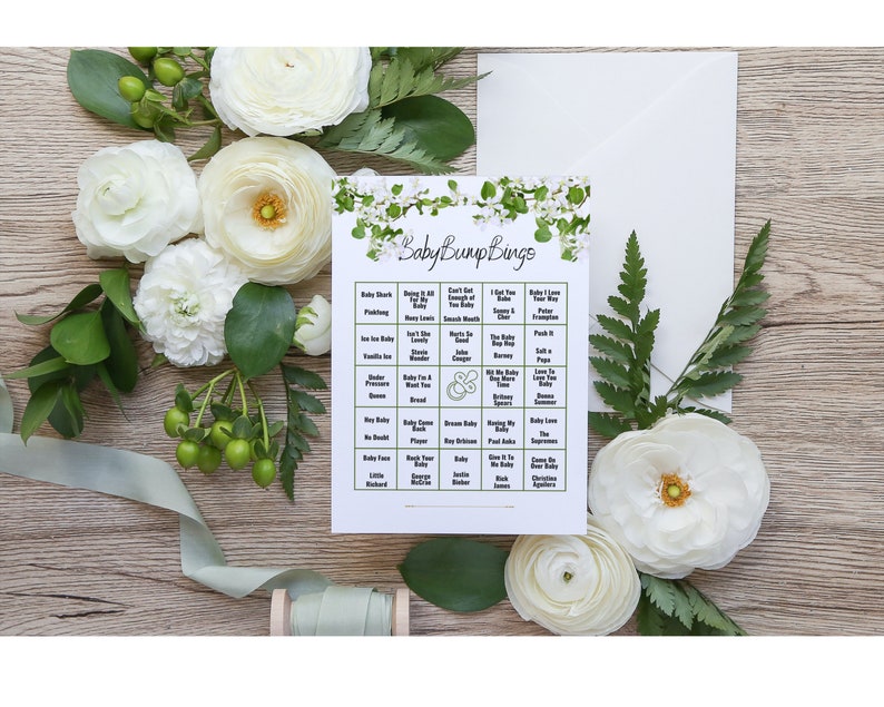 May include: A printable baby shower bingo card with a floral design. The card has a green and white floral border and the text "Baby Bump Bings" at the top. The bingo card has 25 squares with different phrases related to pregnancy and babies.