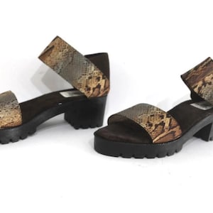 May include: A pair of platform sandals with a snakeskin pattern on the straps. The sandals have a black sole and a block heel. The open-toe design features a wide strap across the foot and an ankle strap.
