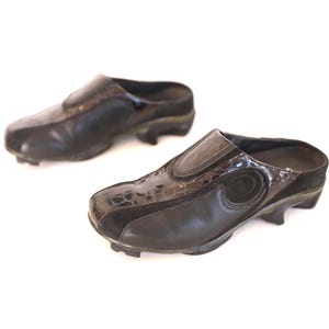 May include: A pair of black leather clogs with a slip-on design. The shoes feature a textured, dark brown accent on the upper and a small heel. The clogs are set against a white background.