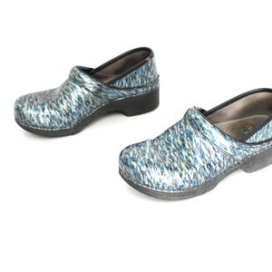 Könnte beinhalten: Ein Paar Clogs mit blauem und silbernem Muster. Die Schuhe haben ein geschlossenes Zehendesign mit niedrigem Absatz und schwarzer Sohle. Die Innenseite des Schuhs ist hellbeige.