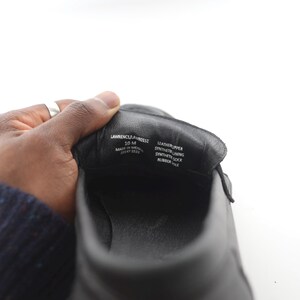 May include: Close-up of the inside of a black leather shoe. The shoe is labeled "Lawrence &  Webster" and "10 M" in white text. The shoe also has the following information printed on the inside: "Leather Upper", "Synthetic Lining", "Synthetic Sock", "Rubber Sole".