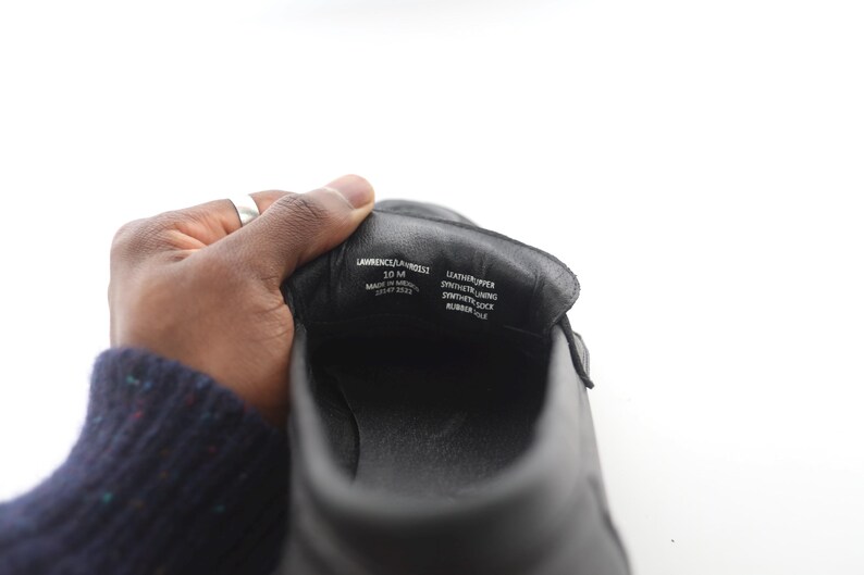 May include: Close-up of the inside of a black leather shoe. The shoe is labeled "Lawrence &  Webster" and "10 M" in white text. The shoe also has the following information printed on the inside: "Leather Upper", "Synthetic Lining", "Synthetic Sock", "Rubber Sole".