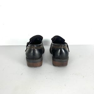 May include: A pair of black leather monk strap shoes featuring a single side buckle closure, a low stacked heel, and visible brown stitching along the heel counter and collar. The shoes have a smooth finish and a rounded toe profile.