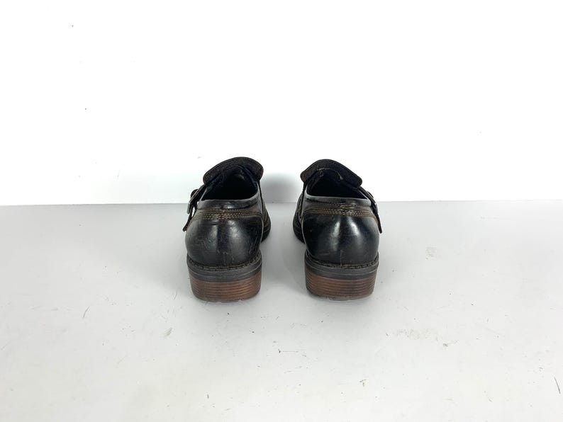 May include: A pair of black leather monk strap shoes featuring a single side buckle closure, a low stacked heel, and visible brown stitching along the heel counter and collar. The shoes have a smooth finish and a rounded toe profile.