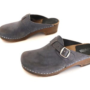May include: A pair of gray suede clogs with wooden soles. Each clog features a buckle closure and white stitching around the sole. The clogs are displayed on a white background.