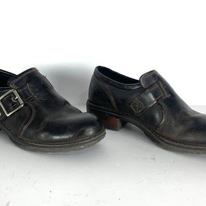 May include: A pair of dark brown leather shoes featuring a rounded toe, a low block heel, and a side strap with a silver-toned metal buckle closure. The shoes have visible contrast stitching along the seams and a textured rubber sole for traction.