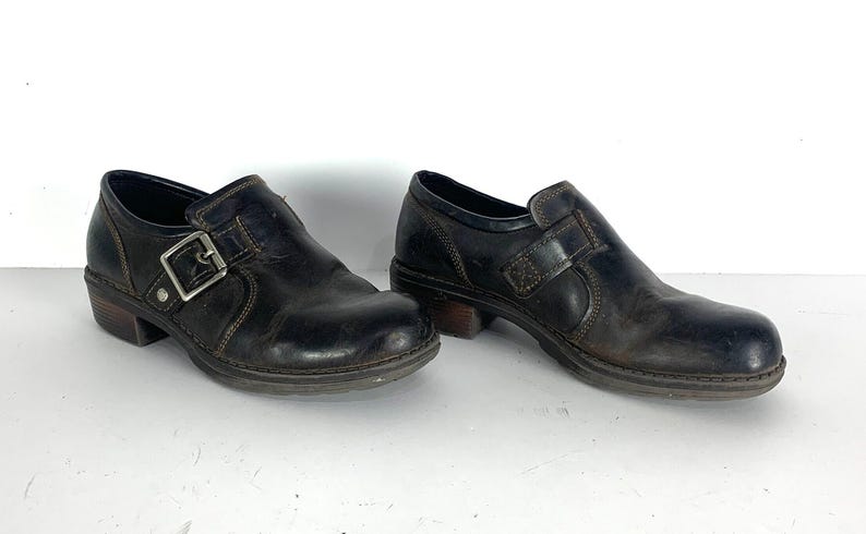 May include: A pair of dark brown leather shoes featuring a rounded toe, a low block heel, and a side strap with a silver-toned metal buckle closure. The shoes have visible contrast stitching along the seams and a textured rubber sole for traction.