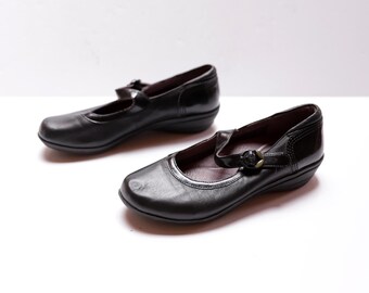 May include: Pair of black leather Mary Jane style shoes with a strap and buckle closure. The shoes have a low heel and a rounded toe. The shoes are set against a white background.