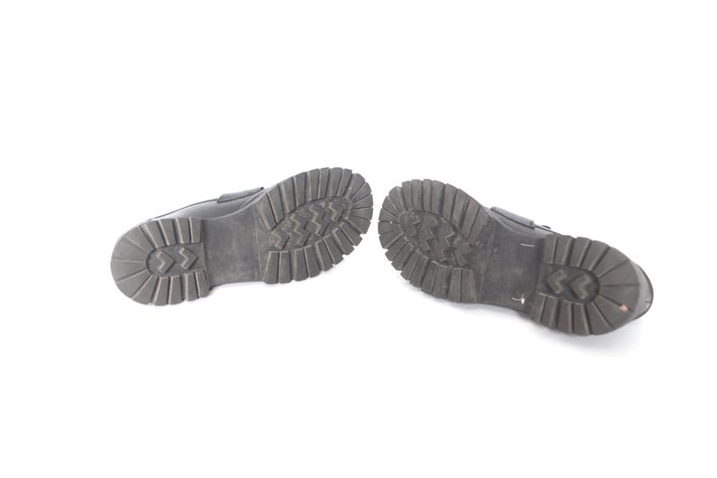 May include: A pair of black leather shoes with a textured rubber sole. The shoes are shown from the bottom, with the soles facing the camera.