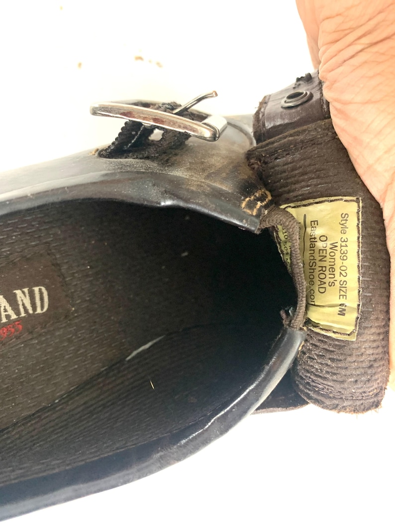 May include: A dark brown leather shoe with a silver-toned metal buckle strap. The interior features a black textured insole with the brand name Eastland. A yellow tag inside displays the text Style 3139-02 SIZE 6M OPEN ROAD EastlandShoe.com.