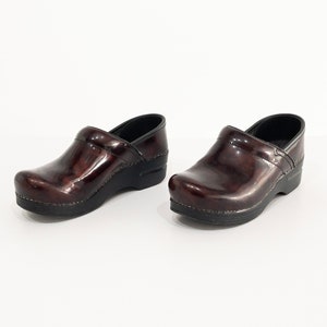May include: A pair of dark brown leather clogs with a glossy finish. The clogs have a black rubber sole and a rounded toe.