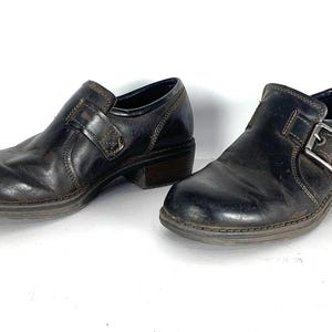May include: A pair of dark brown leather shoes featuring a rounded toe, a low block heel, and a side strap secured with a silver-toned metal buckle. The shoes have visible contrast stitching along the seams and a smooth, polished finish.