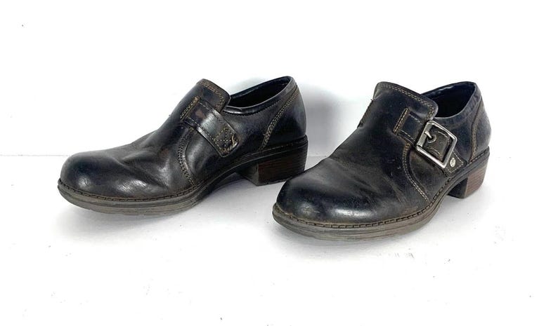 May include: A pair of dark brown leather shoes featuring a rounded toe, a low block heel, and a side strap secured with a silver-toned metal buckle. The shoes have visible contrast stitching along the seams and a smooth, polished finish.