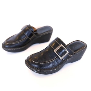 Pode incluir: Um par de tamancos de couro preto com salto cunha baixo. Os tamancos apresentam um fecho de fivela e detalhes de costura brancos. Os sapatos estão sobre um fundo branco.