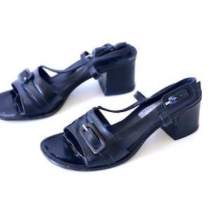 May include: A pair of black leather sandals with a block heel. The sandals have a strap across the toes and a buckle closure on the ankle strap. The heels are approximately 2.5 inches high.