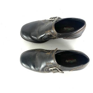 May include: A pair of dark brown leather slip-on shoes featuring a side buckle strap detail. The interior footbed is black and displays the brand name EASTLAND in white text. The shoes have a rounded toe and a low heel profile.