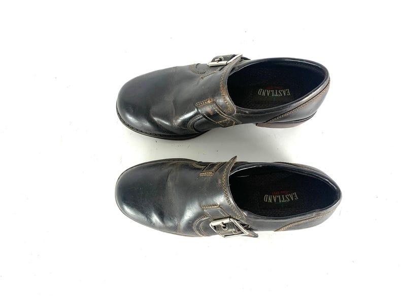 May include: A pair of dark brown leather slip-on shoes featuring a side buckle strap detail. The interior footbed is black and displays the brand name EASTLAND in white text. The shoes have a rounded toe and a low heel profile.