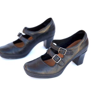 May include: A pair of black leather Mary Jane style shoes with double buckle straps and block heels. The shoes have a rounded toe and a platform sole. The heels are approximately 2.5 inches (6.35 cm) high.