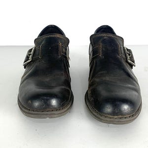 May include: A pair of dark brown leather shoes featuring a rounded toe, a single side strap with a silver-toned metal buckle closure, and visible brown stitching along the edges. The shoes have a low-profile design with a dark sole.