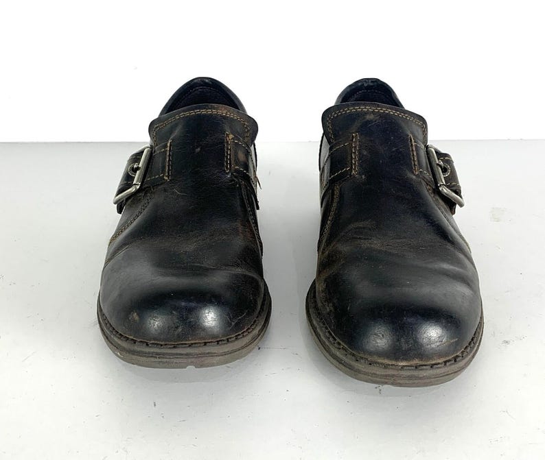 May include: A pair of dark brown leather shoes featuring a rounded toe, a single side strap with a silver-toned metal buckle closure, and visible brown stitching along the edges. The shoes have a low-profile design with a dark sole.