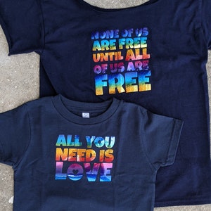 May include: Two navy blue t-shirts with rainbow-colored text. The larger shirt says "None of us are free until all of us are free". The smaller shirt says "All you need is love".
