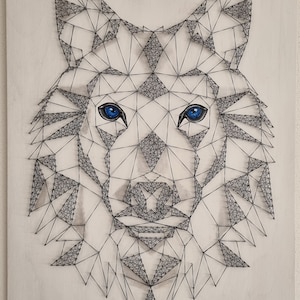 Könnte beinhalten: Ein weißer Wolfskopf wird mit String Art auf einem hellen Holzgrund geschaffen. Die Augen des Wolfes sind blau und die String Art ist schwarz.