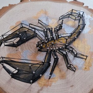 Könnte beinhalten: Ein schwarzer String-Art-Skorpion auf einer Holzscheibe. Der Skorpion ist mit schwarzem Garn umrandet und hat einen gold- und silberfarbenen Hintergrund.