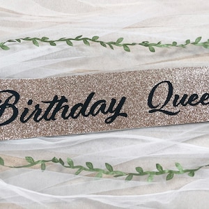 May include: A gold glitter sash with the text "Birthday Queen" in black cursive lettering.