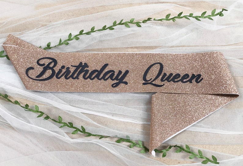 May include: A gold glitter sash with the text "Birthday Queen" in black lettering.