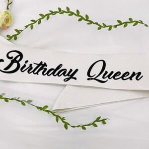May include: White satin sash with black glitter lettering that reads "Birthday Queen".