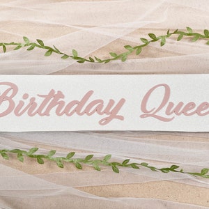 May include: A white banner with the words "Birthday Queen" written in pink cursive letters. The banner is decorated with green leafy vines.